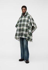 Άνδρας που φοράει πράσινο και λευκό καρό πόντσο με κουκούλα, μπλε τζιν και καφέ παπούτσια, στέκεται μπροστά σε απλό φωτεινό φόντο.