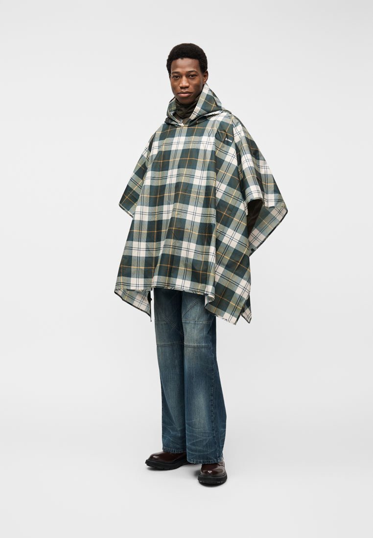 Homme portant un poncho à capuche à carreaux verts et blancs, un jean bleu et des chaussures marron, debout devant un fond clair uni.