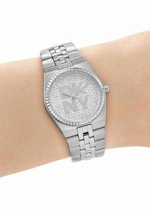 Silberne DKNY-Armbanduhr mit mit Strass besetztem Zifferblatt und Logo, getragen an einem hellhäutigen Handgelenk mit einem Metallgliederarmband.