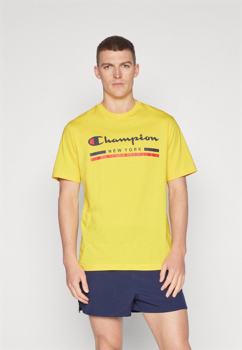 Camiseta de algodón amarilla con un logo en negro y rojo de "Champion New York" en el pecho. Manga corta y un diseño clásico de cuello redondo.