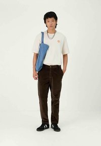 Jovem vestido com uma t-shirt branca com um logótipo do sol, calças castanho-escuro, sapatilhas pretas, correntes de prata e uma mala azul ao ombro, em pé contra um fundo liso.