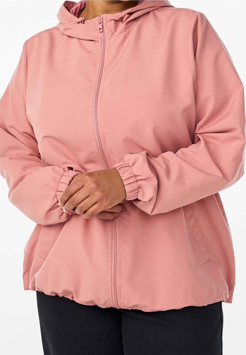 Personne portant une veste à capuche rose clair avec fermeture éclair, poignets élastiques et poches latérales, debout les mains jointes devant elle.