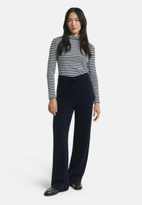 Vrouw in een zwart-witte gestreepte lange mouwtop, marineblauwe wijde broek met trekkoord en donkerbruine schoenen, staand tegen een witte achtergrond.