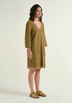 Woman standing wearing a yellow and purple patterned knee-length dress with three-quarter sleeves and matching slippers.