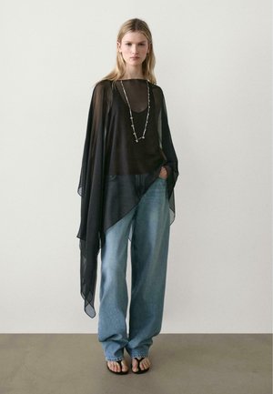 Donna che indossa un top nero trasparente asimmetrico, una lunga collana, jeans blu larghi e sandali neri, in piedi contro una parete chiara e uniforme.