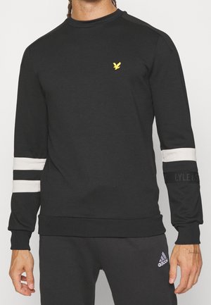 Mężczyzna ubrany w czarną bluzę z białymi paskami na rękawach i małym żółtym logo ptaka na piersi, w połączeniu z czarnymi spodniami Adidas.