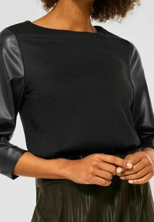 Black top with leather sleeves worn by person with medium skin tone, hands clasped, paired with dark pleated skirt.