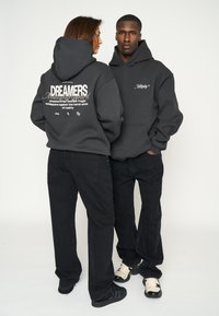 Donkere grijze hoodies hebben contrasterende witte tekst en ontwerpen op de achterkant. Beide modellen dragen zwarte broeken en schoeisel met een gestructureerde afwerking.