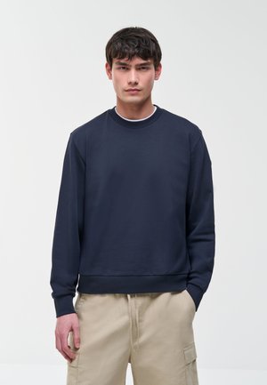 Jonge man met donker haar die een marineblauwe sweatshirt en beige cargobroek draagt, staand tegen een effen lichte achtergrond.