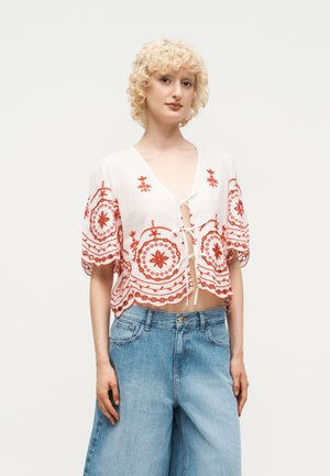 Blond persoon met krullend haar die een witte cropped blouse met rode bloemenborduursel en lichtblauwe jeans met wijde pijpen draagt.