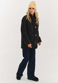 Czarna pikowana kurtka z kolorowymi guzikami, w połączeniu z niebieskimi dzwonowatymi jeansami i żółtą dzianinową czapką. Stylizację dopełniają botki.