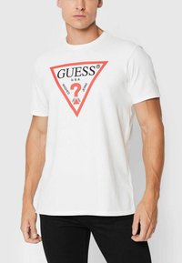 T-shirt bianco a maniche corte con logo triangolare rosso e testo nero "GUESS U.S.A. WASHED JEANS" e un punto interrogativo al centro.