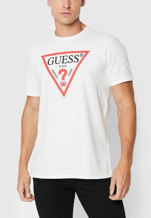 T-shirt bianco a maniche corte con logo triangolare rosso e testo nero "GUESS U.S.A. WASHED JEANS" e un punto interrogativo al centro.