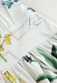 Tissu blanc avec des plis, présentant des illustrations botaniques imprimées de fleurs jaunes, bleues et vertes, et un texte en cursive indiquant "Laven".