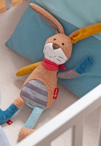Soft stuffed bunny with multicolored limbs, striped belly, red scarf, lying on a bed with blue cushions.