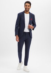 Garnitur w granatową prążkowaną tkaninę o dopasowanym kroju, marynarka na jeden guzik, kieszeń naszywana z czerwonym akcentem, zestawiona z białymi trampkami i białą koszulą.