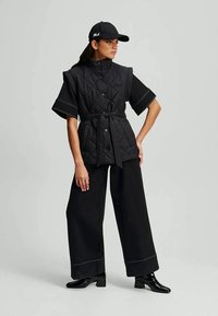 Juoda siuvinėta liemen�ė su trumpomis rankovėmis, surišta juosmens srityje, derinama su plačiomis juodomis kelnėmis. Juoda kepurė su logotipu, blizgūs kulkšnies aulinukai.