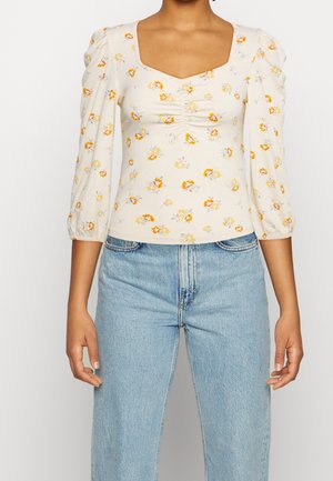 Woman wearing cream long-sleeve top with orange floral print and ruched neckline, paired with light blue high-waisted jeans.
