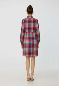 Robe à carreaux rouge et bleu avec de longues manches bouffantes, col, taille cintrée et coupe au niveau du genou. Portée avec des chaussures à talons beiges.