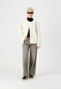 Veste blanche, texturée, de style boxy avec trois boutons, associée à un jean gris à jambes larges et des chaussures noires à semelle légère. Un col roulé noir en dessous.