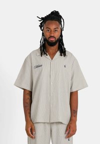 Camisa de manga corta a rayas en beige y blanco, con botones en la parte delantera y logotipos bordados en azul marino. Fabricada con tela ligera.