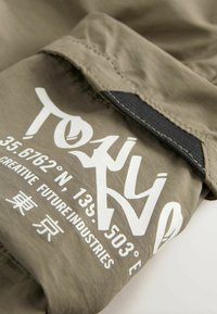 Tessuto khaki con testo bianco che legge "TOKYO" e coordinate. Comprende un accento nero al colletto e dettagli cuciti.