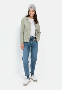 Lichtgroene jas met knopen, witte shirt eronder, blauwe spijkerbroek en witte sneakers met grijze accenten. Een grijze gebreide beanie maakt de look compleet.