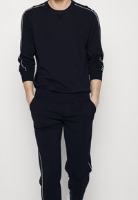 Homme portant un sweat-shirt noir à manches longues et un pantalon assorti avec un liseré blanc, mains dans les poches, debout devant un fond blanc uni.