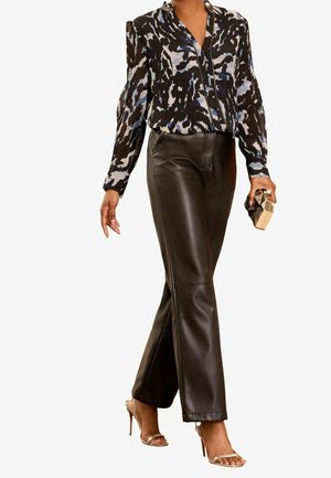 Frau trägt eine schwarze, plissierte Bluse mit Muster, schwarze Lederhose, goldene Sandalen mit hohem Absatz und hält eine geometrische goldene Clutch.