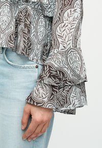 Main reposant à côté d'un jean délavé clair, portant un chemisier à motifs avec des manches superposées et évasées ornées d'un motif de feuilles et de vignes.