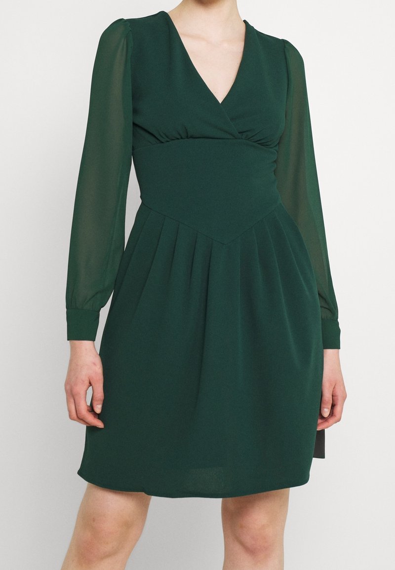 Grön klänning med djup V-ringning, långa genomskinliga ärmar, plisserad kjol och åtsittande midja, tillverkad av lättviktsmaterial.