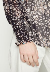 Blusa nera e beige con motivo a fantasia e polsini arricciati, caratterizzata da un design smock. Il materiale sembra leggero e trasparente con accenti blu.