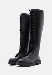 Μαύρες δερμάτινες μπότες μέχρι το γόνατο με σχέδιο δέσιμο μπροστά, ανθεκτικές σόλες και διακριτικούς πλαϊνούς φερμουάρ για επιπλέον λειτουργικότητα.