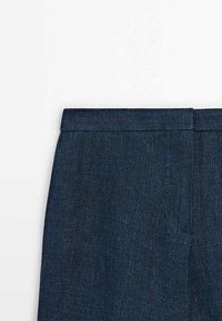Gros plan sur la ceinture d'un pantalon bleu marine sur mesure, montrant le tissu texturé et la couture de fermeture supérieure sur un fond blanc.