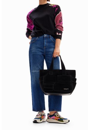 Personne portant un pull noir à fleurs, un jean bleu, des baskets multicolores, tenant un sac fourre-tout noir Desigual avec plusieurs poches.