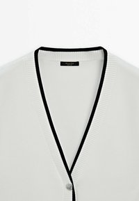 Cardigan blanc à col en V avec une bordure noire le long de l'encolure et de l'ouverture avant, avec un seul bouton visible près du bas.