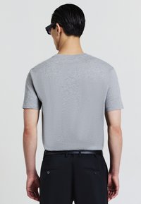 Uomo con capelli neri lisci, che indossa occhiali da sole neri, una camicia grigia a maniche corte e pantaloni neri, mostrato di spalle su uno sfondo neutro.