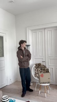 Rudas užtrauktukas megztinis, pilka marškiniai, tamsiai mėlynos kelnės, stovintis ryškiame kambaryje. Balta kėdė su kailio užvalkalu, "Zalando" dėžė ant mažo medinio staliuko.