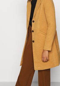 Mustard yellow wool blend coat with a single-breasted design, three buttons, and side pockets, paired with brown pants.