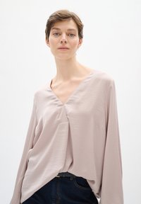 Blusa di un leggero color malva con scollatura a V, maniche lunghe e vestibilità rilassata. Tessuto liscio con una leggera lucentezza, dotata di un orlo asimmetrico.