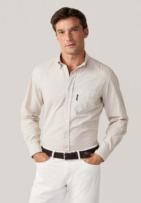 Camicia button-up a maniche lunghe in cotone beige chiaro con taschino sul petto, abbinata a pantaloni bianchi e una cintura intrecciata marrone.