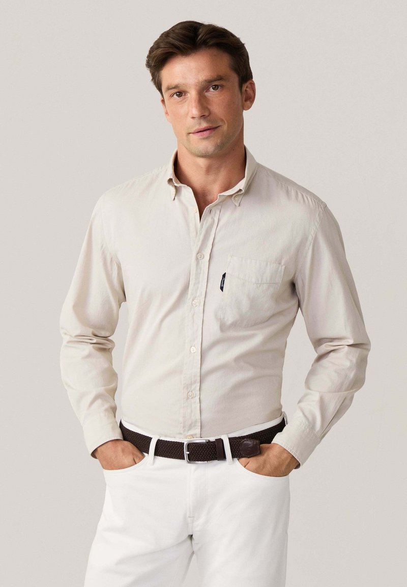 Camicia button-up a maniche lunghe in cotone beige chiaro con taschino sul petto, abbinata a pantaloni bianchi e una cintura intrecciata marrone.