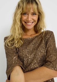Top marrón con lentejuelas, de corte holgado, escote redondo y mangas cortas. La textura es brillante y Reflectante, creando un diseño llamativo.