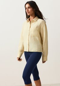Femme portant une chemise jaune rayée à boutons et des leggings bleu marine courts, debout les bras détendus.