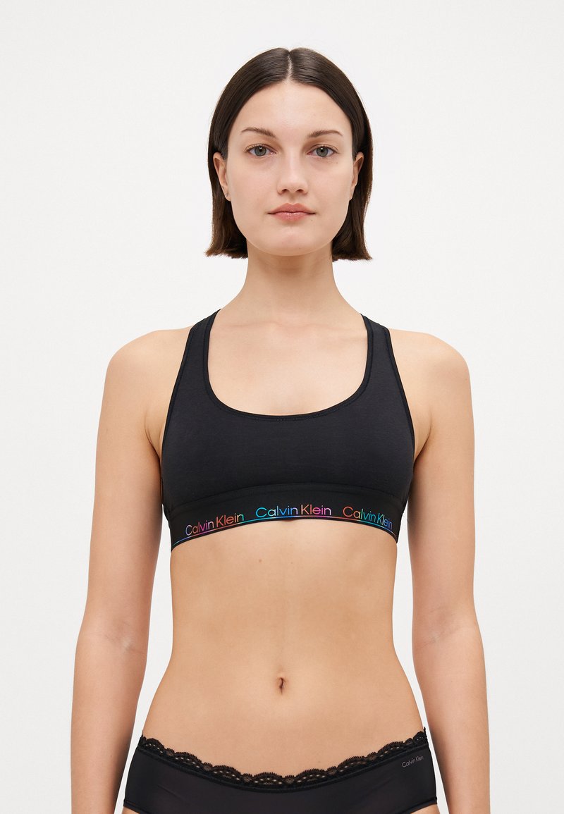 Svart sportsbh laget av myk stoff, med en bred kant som har flerfarget "Calvin Klein"-logo. Enkel design, racerback-stil.