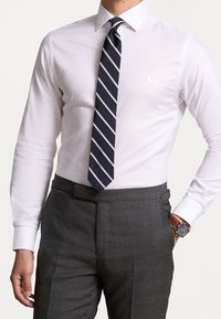 Biała koszula z długimi rękawami, połączona z granatowym, paskowanym krawatem. Ciemnoszare, dopasowane spodnie z eleganckim zegarkiem na nadgarstku.