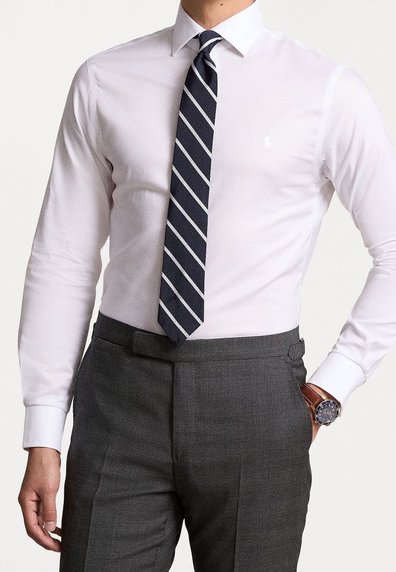 Biała koszula z długimi rękawami, połączona z granatowym, paskowanym krawatem. Ciemnoszare, dopasowane spodnie z eleganckim zegarkiem na nadgarstku.