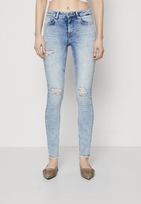 Light blue distressed skinny jeans with ripped detailing, five-pocket design, and a fitted silhouette. Paired with brown flat shoes.