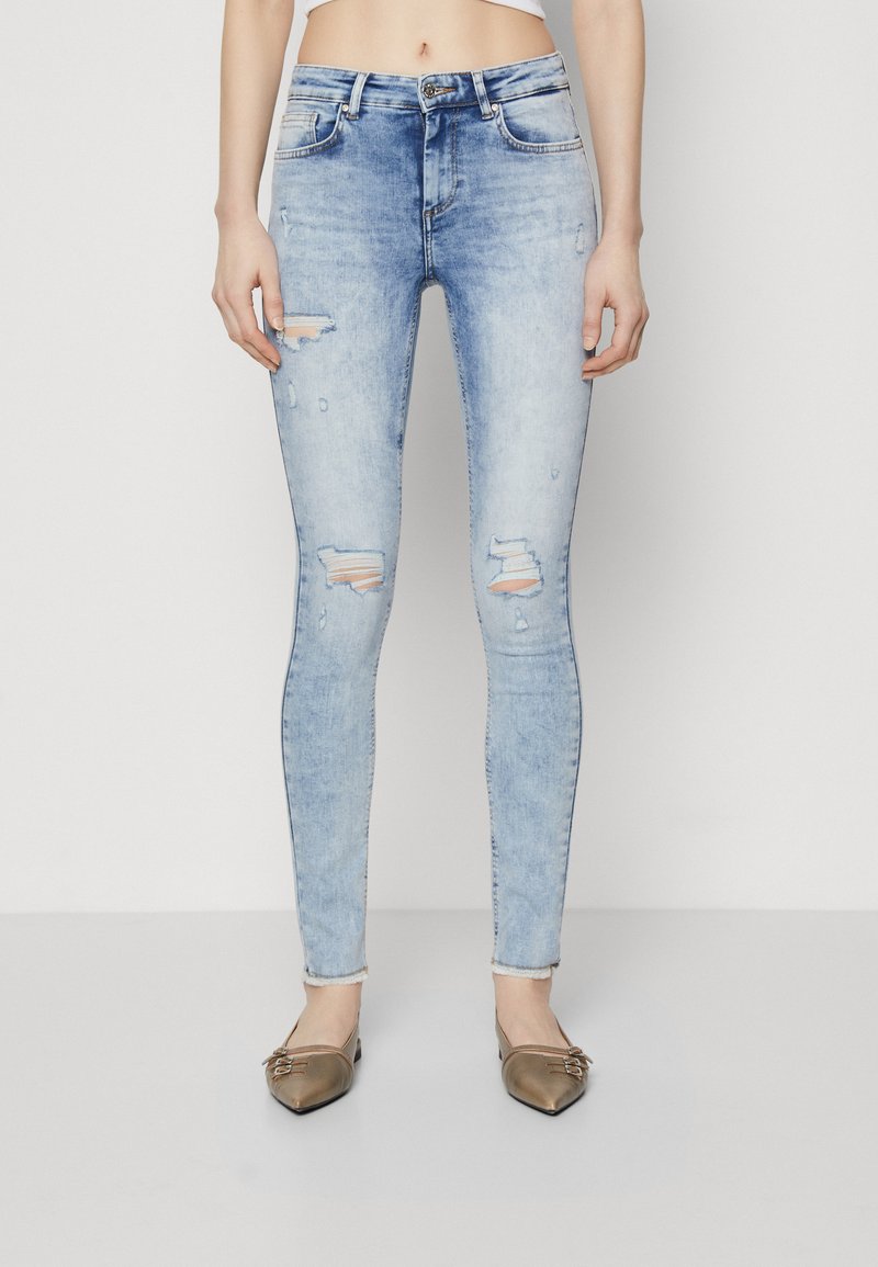 Light blue distressed skinny jeans with ripped detailing, five-pocket design, and a fitted silhouette. Paired with brown flat shoes.