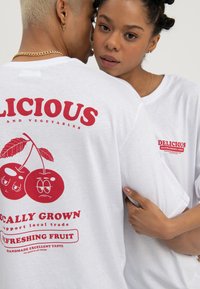 Femme embrassant un homme par derrière, tous deux portant des t-shirts blancs avec des imprimés fruités rouges "Délicieux", la femme regardant directement la caméra.